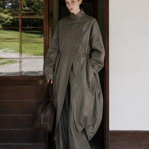 Könnte beinhalten: Ein olivgrüner langer Mantel mit Stehkragen, asymmetrischer Knopfleiste und Kordelzug an der Taille, kombiniert mit einer passenden weiten Hose und einer dunkelbraunen Ledertasche mit silbernem Reißverschluss und zwei Tragegriffen.