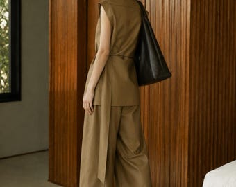 May include: A sleeveless olive-brown tunic top with a wrap-around tie waist and matching wide-leg trousers. The outfit is paired with a large black leather shoulder bag and pointed-toe black shoes, creating a coordinated monochromatic ensemble.