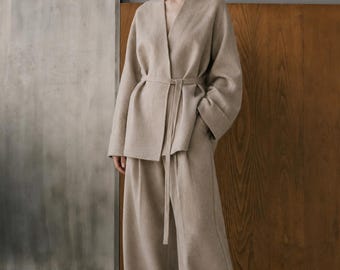 May include: A beige two-piece set consisting of a long-sleeved wrap jacket with a tie waist and wide-leg trousers. The jacket features a V-neckline and loose sleeves, while the trousers have a relaxed fit. The fabric has a soft, felted texture.