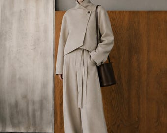 May include: A beige wool-blend ensemble consisting of a cropped jacket with a high collar and single button closure, paired with high-waisted wide-leg trousers featuring a matching tie belt. A dark brown cylindrical leather shoulder bag is worn.