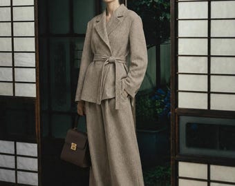 May include: A beige textured blazer with a notched lapel and a matching tie-waist belt, paired with wide-leg trousers in the same fabric. A structured dark brown leather handbag with a gold-tone clasp is held at the side.