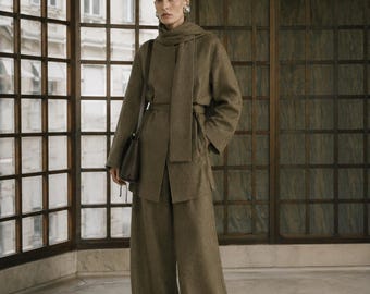 May include: An olive green wool-blend ensemble consisting of a long-sleeved jacket with a matching scarf draped over the shoulders, a belted waist, and wide-leg trousers. A dark brown leather shoulder bag is carried on the side of the jacket.