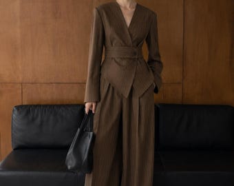 May include: A brown pinstriped blazer with a V-neckline and a wide integrated waist belt, paired with matching wide-leg trousers. The blazer features long sleeves and a single button closure at the waist. A black leather handbag is held at the side.