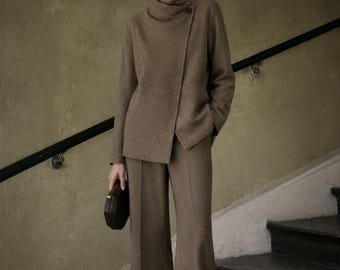 May include: A taupe two-piece suit consisting of a long-sleeved jacket with a high collar, an asymmetrical button closure, and matching wide-leg trousers with a center crease, paired with a small dark brown rectangular clutch bag.