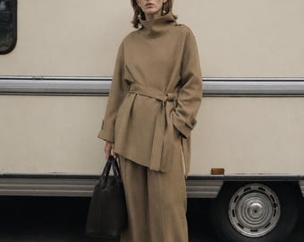 May include: A tan high-neck tunic with a matching waist tie and long sleeves, paired with wide-leg trousers in the same tan color. The outfit is completed by a dark brown structured handbag with two top handles and pointed-toe black shoes.