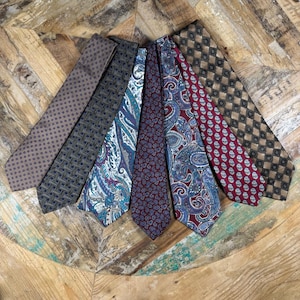 Könnte beinhalten: Eine Sammlung von sieben Krawatten, die fächerförmig angeordnet sind. Die Krawatten weisen verschiedene Muster auf, darunter kleine Punkte, geometrische Quadrate und komplexe Paisley-Designs in den Farben Braun, Blau, Türkis, Burgunderrot, Rot und Beige.