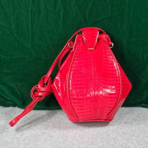 Puede incluir: Un bolso de mano rojo brillante con un patrón texturizado de piel de serpiente y un acabado brillante. El bolso presenta una forma estructurada de diamante con un cierre de solapa superior y una correa larga y fina con un nudo.