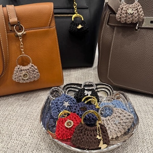 May include: A collection of miniature crochet handbag charms in various colors including red, brown, black, blue, and beige, each featuring a gold or silver tone handle and a decorative metal charm, displayed in a glass bowl and on larger handbags.