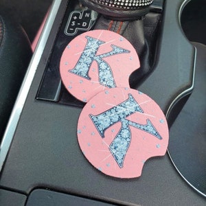 May include: Two pink car coasters with a rhinestone letter K on each. The coasters are in a car cup holder. A black leather gear shift knob with a rhinestone crown is in the background.