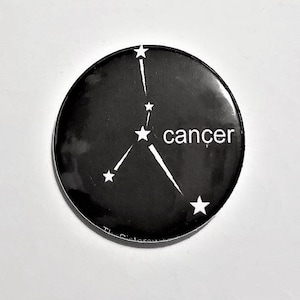 Puede incluir: Botón negro y blanco con un contorno blanco de la constelación de Cáncer y la palabra "cáncer" en texto blanco.