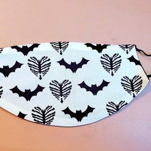 May include: A white face mask with a black and white pattern of bats and ribcages in the shape of hearts. The mask has black elastic ear loops.