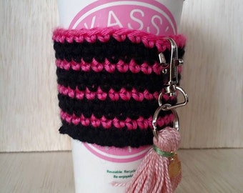May include: A pink and black crocheted coffee cup cozy with a pink tassel attached to a silver key chain. The cozy is on a white to-go coffee cup with the word "VASS" printed on the side.