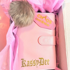 Könnte beinhalten: Ein rosa Lederplaner mit einer goldenen Inschrift, die "Kassy Dee" lautet, und einem rosa Tab, auf dem "Chaos Coordinator" steht. Der Planer befindet sich in einer rosa Geschenkbox mit Seidenpapier.