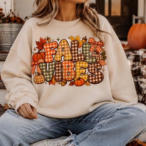 Peut inclure: Un sweat-shirt à col rond beige avec une grande impression graphique portant le texte FALL VIBES dans un motif à carreaux multicolore. Le design est orné d'illustrations de citrouilles orange, de feuilles d'automne rouges et jaunes, de glands et de nœuds.