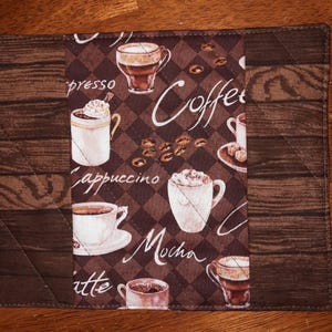 May include: A rectangular quilted fabric mat featuring a central panel with a brown diamond pattern, coffee bean illustrations, and various coffee cups. The text reads Coffee, Espresso, Cappuccino, Mocha, and Latte. The side panels have a wood grain print.