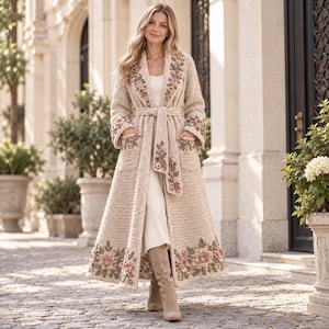 May include: A long, beige, crocheted duster coat featuring a shawl collar, long sleeves, and a tie-waist belt. The garment is decorated with intricate, three-dimensional floral embroidery in shades of pink and green along the hem, pockets, and lapels.