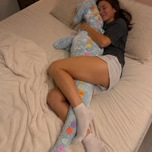 May include: A long, plush body pillow shaped like a dinosaur, featuring a light blue textured fabric with a row of colorful, circular felt patches in orange, yellow, purple, and pink running down the center of its back and tail.