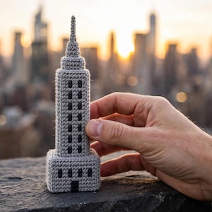 May include: A grey crocheted model of a skyscraper featuring a tiered structure, a pointed spire, and dark grey rectangular accents representing windows. The textile construction shows visible yarn stitches throughout the entire surface of the building.