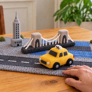 May include: A crochet play mat featuring a gray road with white dashed lines, a yellow taxi, a gray skyscraper, and a beige suspension bridge. The mat includes small green crochet bushes and a blue rectangular section representing water.