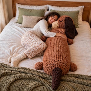 May include: A large, long, brown crocheted dachshund plush toy with dark brown ears and a dark brown tail. The stuffed animal features a textured yarn construction, a rounded snout, and closed eyes, lying horizontally across a white bedspread.