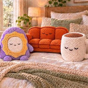 May include: Three crocheted plush items: a purple sun-shaped figure with a yellow border, an orange three-seat sofa, and a white coffee mug with a brown top. Each item features a simple embroidered face with closed eyes and a small smile.