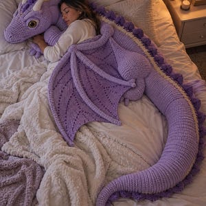 May include: A large, lavender-coloured crochet dragon plush toy featuring a long, segmented tail, detailed wings with open-work stitching, white horns, and large, expressive eyes. The dragon is crafted with a textured yarn finish throughout.