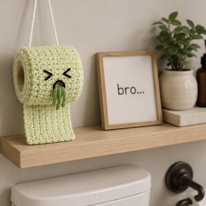 May include: A light green crocheted toilet paper roll with a hanging string, featuring a stitched face with closed eyes and a mouth expelling green yarn. Next to it is a wooden frame containing the text "bro..." and a small potted green plant.
