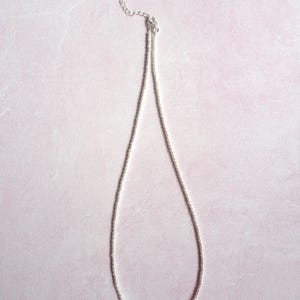 以下が含まれることがあります： 小さな白いビーズが一列に並んだ繊細なネックレス。上部にはシルバーカラーの金属製チェーンエクステンションが付いており、小さな円形のタグで留められています。チェーンにより長さを調節することが可能です。