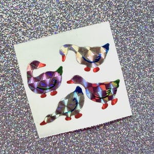 May include: A set of four holographic stickers featuring duck shapes on a white square backing. Each duck has a geometric, multi-faceted surface that reflects light in various colors, with distinct red beaks and feet, and dark blue wing details.