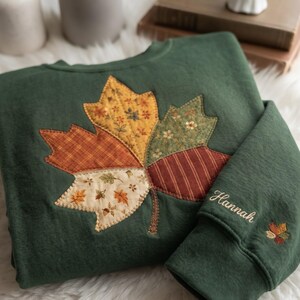 May include: A dark green sweatshirt featuring a large, quilted maple leaf appliqué on the front, composed of various fabric patterns in shades of orange, yellow, and cream. The sleeve is embroidered with the name Hannah and a small leaf icon.