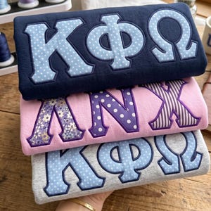May include: A stack of three folded sweatshirts featuring Greek letter appliqués. The top navy sweatshirt has light blue polka dot letters, the middle pink sweatshirt has floral and polka dot letters, and the bottom grey sweatshirt has light blue polka dot letters.