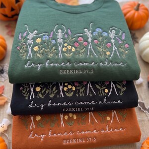 May include: A stack of three crewneck sweatshirts in dark green, black, and burnt orange. Each features embroidered skeletons dancing among colorful wildflowers with the white text "dry bones come alive" and "EZEKIEL 37:5" stitched below.