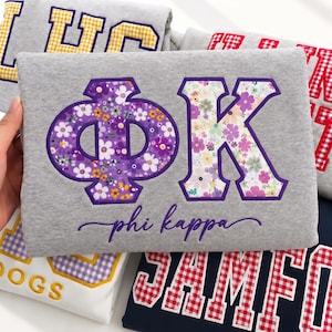 Könnte beinhalten: Ein graumeliertes Sweatshirt mit den griechischen Buchstaben Phi Kappa in einem Blumenmuster mit violetten Umrandungen. Der Text phi kappa ist in violetter Schreibschrift unter den Buchstaben eingestickt. Weitere Sweatshirts sind sichtbar.