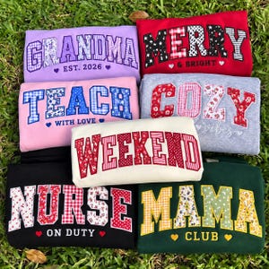 May include: A collection of seven folded sweatshirts in various colours, each featuring large block-letter text with patterned fabric appliques. The shirts display phrases including Grandma Est. 2026, Merry & Bright, Teach With Love, Cozy Vibes, Weekend, Nurse On Duty, and Mama Club.