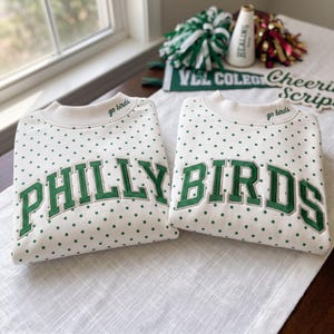 Könnte beinhalten: Zwei cremeweiße Sweatshirts mit Rundhalsausschnitt und grünem Punktemuster. Jedes Sweatshirt zeigt den Schriftzug "PHILLY BIRDS" in großen, grünen, gestickten Blockbuchstaben auf der Brust und den Text "go birds" am Kragen gestickt.