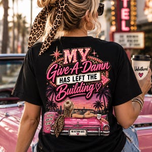 以下が含まれることがあります： 背面にグラフィックプリントが施された黒いTシャツ。デザインには、ピンクと白の文字で「MY Give-A-Damn HAS LEFT THE BUILDING」、ピンクのオープンカー、ピンクのスーツケース、夕日を背景にしたヤシの木が描かれています。