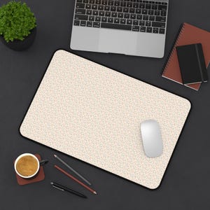 May include: A rectangular desk mat featuring a light beige background with a dense, repeating pattern of tiny multicoloured dots in shades of blue, orange, and yellow, finished with a black stitched border along the entire outer edge.