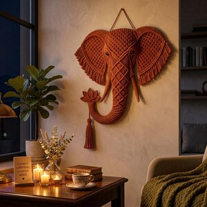 May include: A burnt orange macrame wall hanging shaped like an elephant head with large ears, a trunk, and a lotus flower detail. The piece features intricate knotted patterns and fringe along the ears, suspended by a cord against a wall.