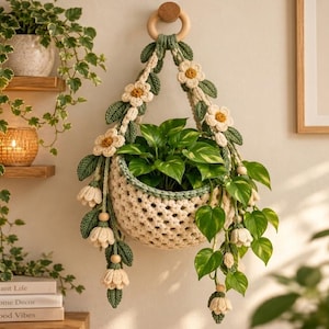 May include: A cream-colored crocheted hanging plant basket featuring green crocheted leaves and white flowers with yellow centers. The basket is suspended from a wooden ring and contains a green leafy plant with trailing vines.