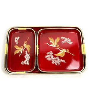 May include: A set of three rectangular red serving trays with gold-toned metal rims and light yellow woven accents on the handles. Each tray features a gold and white graphic illustration of flying cranes and pine trees on a glossy red surface.