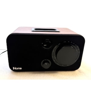 May include: An iHome radio with a wood-grain top and a black fabric front panel. The device features a large circular tuning dial, a smaller volume knob, and a band selector knob. The iHome logo is printed in white on the bottom left corner.