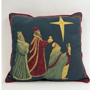 May include: A square throw pillow with a dark teal background featuring a woven tapestry design of three figures in robes and hats, one holding a gold vessel, another a gold jar, beneath a large yellow star, finished with red piping.