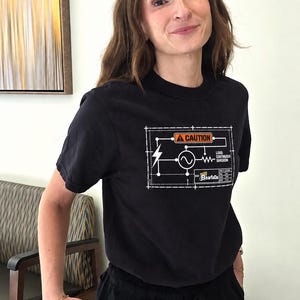 May include: A black short-sleeved t-shirt featuring a white and orange graphic print of an electrical circuit diagram. The design includes a caution symbol, a lightning bolt, and the text "LOAD: CONTINUOUS SARCASM" along with "Beartada".
