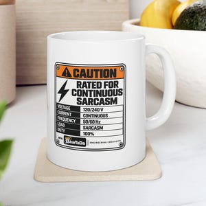 Peut inclure: Un mug en céramique blanc orné d'une étiquette d'avertissement rectangulaire noire et orange. Le texte indique CAUTION RATED FOR CONTINUOUS SARCASM avec des spécifications techniques incluant tension, courant, fréquence, charge et cycle.