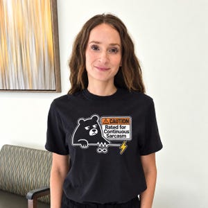 May include: A black t-shirt featuring a graphic of a grumpy bear holding a sign that reads CAUTION Rated for Continuous Sarcasm. The design includes an infinity symbol and a yellow lightning bolt icon, printed in white, orange, and yellow ink.