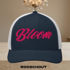 Puede incluir: Una gorra de béisbol azul marino con parte trasera de malla blanca y visera curva. El panel frontal presenta la palabra Bloom bordada en una fuente cursiva de color rosa brillante. La gorra tiene una corona estructurada y un botón.