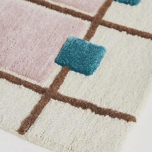 A close-up angled view of the tufted wool rug showing the thick cream base, brown grid lines, pale mauve square, and raised teal square intersection.