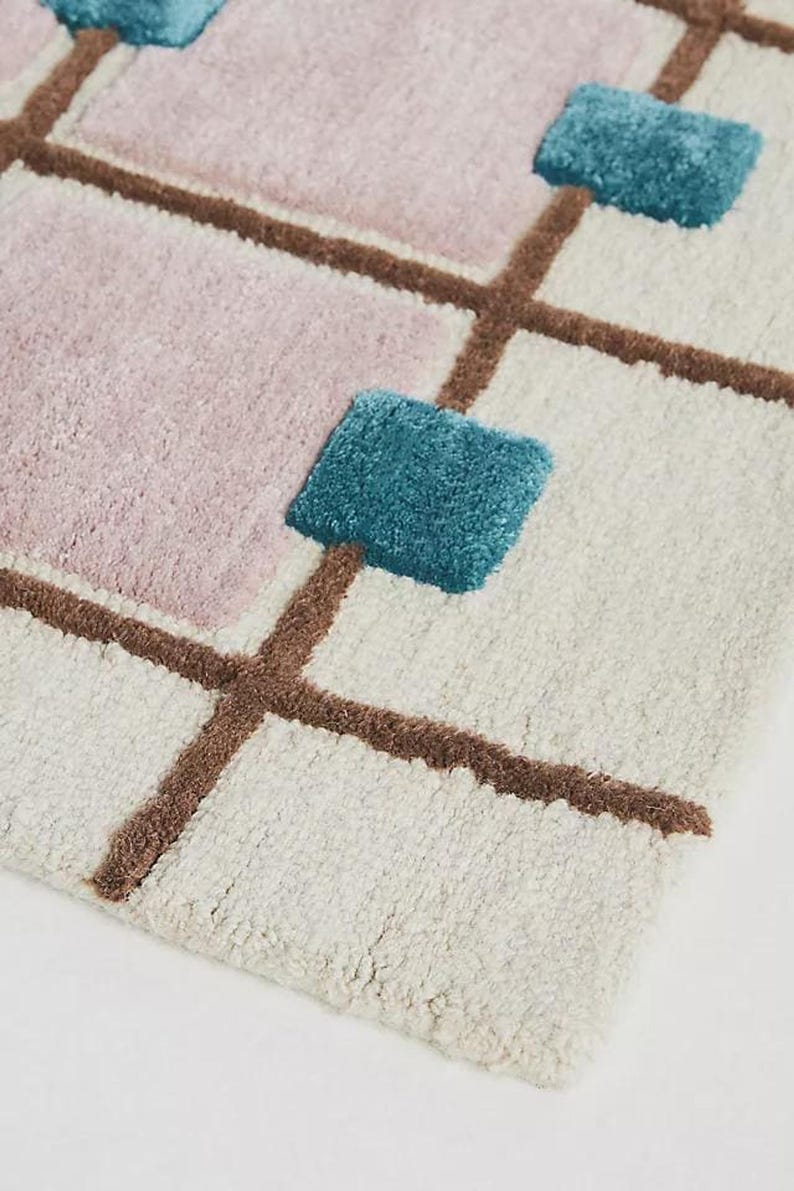 A close-up angled view of the tufted wool rug showing the thick cream base, brown grid lines, pale mauve square, and raised teal square intersection.