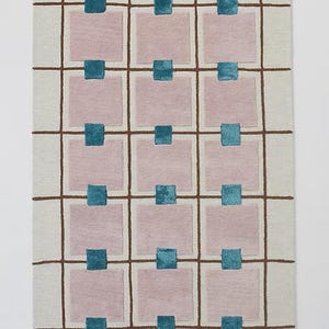 A flat view of a rectangular hand-tufted wool rug featuring an off-white background with a brown grid pattern, mauve squares, and small teal square accents.