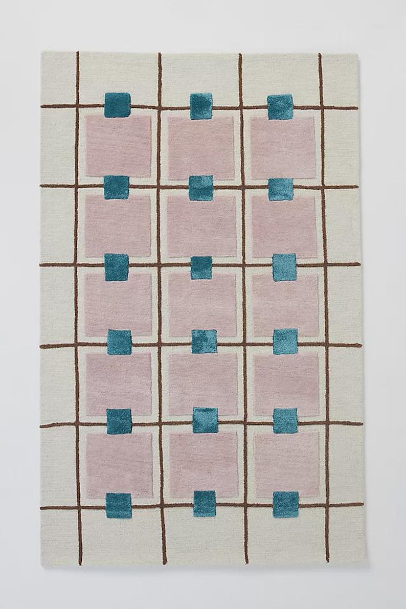 A flat view of a rectangular hand-tufted wool rug featuring an off-white background with a brown grid pattern, mauve squares, and small teal square accents.
